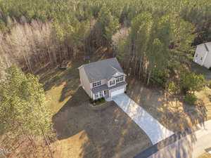 Aerial view of a two-story house with a gray roof and a white garage door, surrounded by a spacious lawn. The property is bordered by a dense forest. A trampoline is visible in the backyard. A long driveway connects the house to a paved road.
