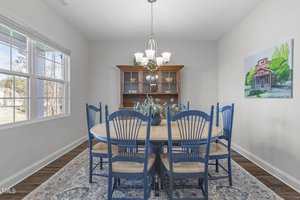 Dining room with a wooden table and six blue chairs. A cabinet with glass doors stands against the wall. A painting of a house hangs nearby. A vase with flowers rests in the center of the table. Large windows let in natural light.