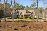 Nestled at 1501 Anterra Drive in Wake Forest, this beautiful, large house features stone and beige siding. Surrounded by bare trees and a landscaped lawn, it boasts dormer windows on the roof and a central double front door, with a paved driveway gracefully curving around it.
