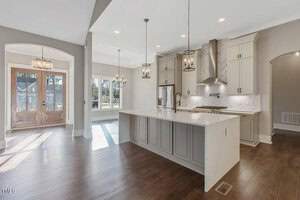 A modern kitchen and dining area with hardwood floors. The kitchen features light gray cabinets, a large island with a white countertop, stainless steel appliances, and two pendant lights. The dining area has large windows and wooden double doors.