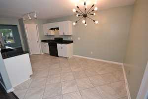 A modern kitchen with light green walls, black granite countertops, white cabinets, and a black stove. A unique chandelier with exposed bulbs illuminates the room. The floor is tiled in a beige pattern. A door leads to an adjacent room.
