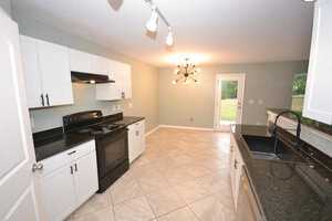 A modern kitchen with white cabinets, black countertops, and tile flooring. It features a black stove and hood, a sink, and a contemporary ceiling light fixture. There is a window and a glass door leading outside, allowing natural light to enter.
