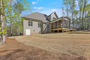 A modern grey house with a steep roof, surrounded by trees, features a screened porch and deck. The spacious, sloping yard is covered in straw and pine needles, set under a clear blue sky.