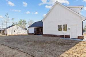 A new white house with a brick foundation is shown from the side in a yard with newly laid sod. The yard is mostly empty and bordered by a wooden fence. A tall fence is visible on the left, with a few houses in the background.