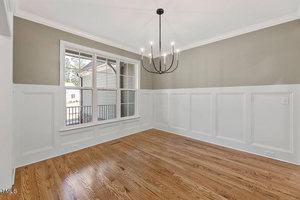 A bright, empty room with light wood flooring and beige walls. It features a large window with an outdoor view and a modern black chandelier hanging from the ceiling. White wainscoting decorates the lower half of the walls.