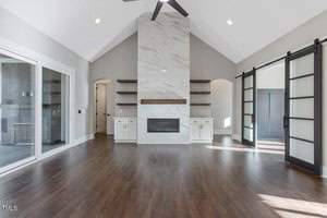 Spacious living room with vaulted ceiling and dark wood floors. Features a modern white fireplace with shelves, large sliding doors, and abundant natural light. White cabinets on either side of the fireplace and glass sliding doors to the sides.