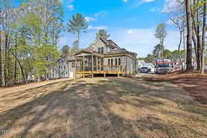 A two-story house with a screened porch sits on a spacious grassy yard surrounded by trees. Nearby, there are other houses and a parked truck on the driveway. The sky is clear and blue.