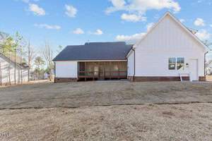A back view of a white house with a dark shingle roof, featuring a screened porch. The lawn is sparse with dry grass, and the sky is partly cloudy. Surrounding trees in the distance add to the suburban setting.
