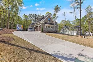 A modern two-story house with a sloped driveway is surrounded by trees. A moving truck and a parked car are in the driveway. The sky is clear, and the grass appears neatly trimmed.
