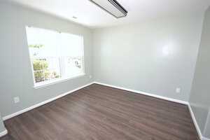 An empty room with pale green walls, a large window with a view of greenery, and dark wooden flooring. The ceiling has a fluorescent light fixture. The room is bright with natural light.