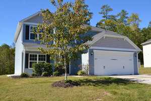 A two-story suburban house with light blue siding and a white garage door. There's a small tree in the front yard and a manicured lawn. The sky is clear and sunny.