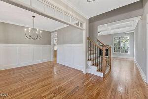 Interior of a house featuring a spacious entryway with polished wooden floors, a staircase with wooden railing, and a glimpse into another room with a large window. White wainscoting and a modern chandelier enhance the elegant decor.