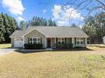 A single-story green house with white trim graces 3044 Cannady Mill Road in Franklinton. Nestled in front of a wooded area, it features a garage on the left, a welcoming front porch with a red door, and a large window. The yard is grassy under a clear sky dotted with a few clouds.