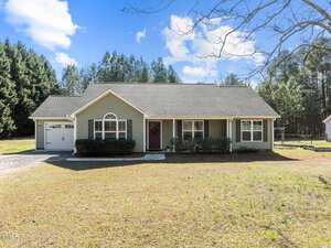 A single-story green house with white trim graces 3044 Cannady Mill Road in Franklinton. Nestled in front of a wooded area, it features a garage on the left, a welcoming front porch with a red door, and a large window. The yard is grassy under a clear sky dotted with a few clouds.
