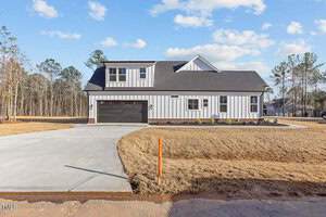A modern two-story white house with black accents and a dark garage door, surrounded by a dry grass lawn and a newly paved driveway. There are trees and a blue sky with clouds in the background.