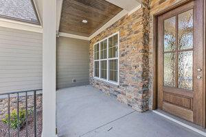 A house entrance with a concrete porch, stone and wooden walls, and a wood-framed glass door. A large window is on the side, and a metal railing runs along the edge of the porch. The ceiling is finished with wooden panels.