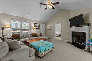 A cozy living room with a vaulted ceiling and fan. It features a large L-shaped sectional sofa with various cushions, a wooden coffee table, a fireplace, and a TV. The room is carpeted and has windows letting in natural light. A bedroom is visible through an open door.