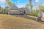 A two-story modern farmhouse at 988 Browning Place in Youngsville features a stone foundation, gray siding, and a gabled roof. Nestled on a sloped, dry grass lawn and surrounded by trees under a blue sky, it includes a few steps leading to its wooden front door.