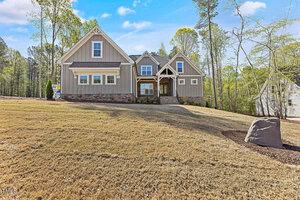 A two-story modern farmhouse at 988 Browning Place in Youngsville features a stone foundation, gray siding, and a gabled roof. Nestled on a sloped, dry grass lawn and surrounded by trees under a blue sky, it includes a few steps leading to its wooden front door.