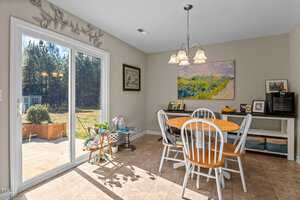 A dining area with a round wooden table and four white chairs. A colorful painting hangs on the wall. Sunlight streams in through a glass sliding door, revealing a view of a backyard with trees. A side table holds plants and decorative items.