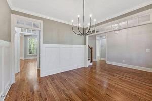 Bright, empty room with polished hardwood floors and light gray walls. Features a black, modern chandelier, large white moldings, and visible stair banister. Sunlit hallway leads to another room in the background.