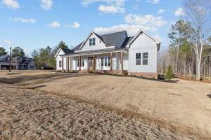 A modern suburban house with a light gray exterior and brick foundation, set against a background of trees and a clear blue sky. The yard in the foreground is freshly landscaped.