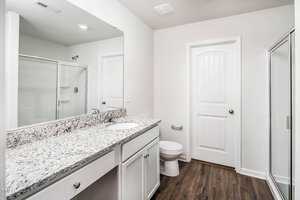 A modern bathroom with white cabinets, granite countertop, and a large mirror above the sink. There is a toilet beside a door and a glass-enclosed shower on the opposite side. The flooring is wood-style laminate.