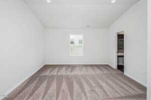 Minimalist empty room with white walls, beige carpet, and recessed lighting. A window on the back wall overlooks a grassy area. An open door on the right reveals a glimpse of a bathroom.