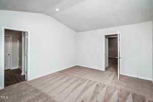 An empty room with white walls and beige carpet, featuring a small closet with an open door on one side. There is an open doorway leading to another room with dark wood flooring. The ceiling is vaulted with a single recessed light.