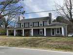 The two-story gray house at 111 W Tally Ho Road, featuring white columns and shutters, sits gracefully on a grassy lawn. With two large chimneys and bare trees surrounding it, a road runs in front as the sky remains partly cloudy.