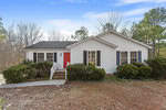 Nestled at 1694 Cobblestone Drive in Creedmoor, this charming white single-story house features a striking red front door and black shutters. A quaint front porch with steps is surrounded by lush bushes and trees, all under a clear blue sky.