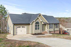A single-story beige house with a dark gray roof and an attached garage. The front features a small porch with stairs and a large arched window. The driveway is concrete and slopes downwards. A red car is parked on the side.