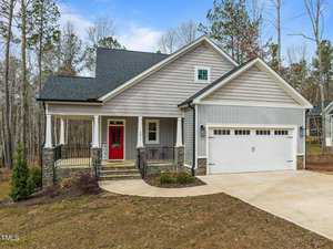 Located at 3611 River Watch Lane, this charming single-story house in Franklinton showcases a gray facade with stone accents. It features a covered front porch with columns, a striking red door, and a double garage, all beautifully set on a landscaped lot with trees providing a serene backdrop.