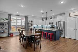Modern kitchen and dining area with hardwood floors. Features a wooden dining table with six chairs, a kitchen island with red stools, white cabinets, stainless steel appliances, pendant lights, and large windows providing natural light.