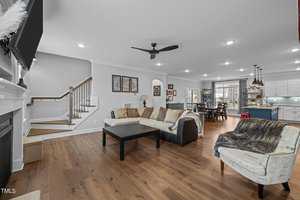 A spacious, modern living room with light wood flooring, featuring a sectional sofa, coffee table, and a TV above a fireplace. A dining area is visible in the background, along with an open kitchen and a staircase to the side.