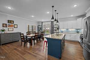 A modern kitchen and dining area with wooden floors. It features a central island with a granite countertop, pendant lights, a dining table with chairs, white cabinets, and a large window overlooking a backyard. Plants and pictures adorn the room.