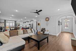 A modern open-plan living room with beige sectional sofa and black coffee table. The kitchen is visible in the background with white cabinets and dining table. The walls are adorned with a clock and artwork. A ceiling fan and wooden flooring complete the space.