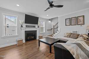 A modern living room with a black ceiling fan, wall-mounted TV, and fireplace. Large windows with blinds brighten the room. There's a black coffee table, beige sectional sofa, and framed art on the walls. A staircase is visible in the background.