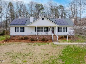 Located at 3536 Fletchers Way, this charming single-story white house features a welcoming front porch and eco-friendly solar panels on the roof. The yard, with sparse grass and bare trees, leads to the porch via a paved path, while a chimney stands proudly on the left.