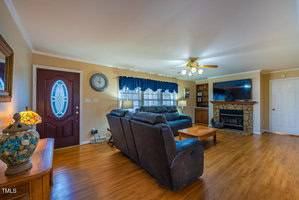 Cozy living room with wooden floor, dark leather sofas, and a stone fireplace. A large TV is mounted on the wall. A round clock hangs above a window with blue curtains. A decorative lamp and a round mirror adorn a table near the entrance.