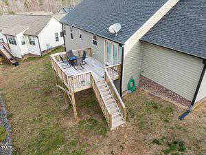 Aerial view of a house with a wooden deck featuring outdoor furniture, including a table and chairs. The deck is connected to the ground by a staircase. The house has beige siding, a gray roof, and is surrounded by a grassy yard. Adjacent structures are visible.