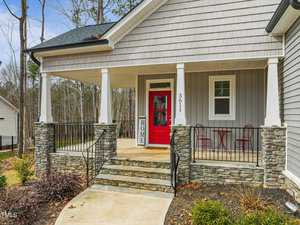 Front porch of a house with a red door and gray siding. The porch features stone columns, a small table with two chairs, and a vertical "HOME" sign. A curved concrete path leads up to the porch, with woods visible in the background.