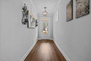 A narrow hallway with wooden floors leads to a white door with a window. Walls are decorated with a map and two framed prints. A lantern-style light hangs from the ceiling, and a potted plant is mounted on the wall. Natural light streams in.