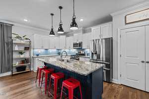 A modern kitchen with white cabinets and a granite island. Three black pendant lights hang above the island, which is lined with red bar stools. Stainless steel appliances are visible, and a bookshelf stands to the side with decor items.