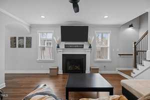 Modern living room with a black coffee table, a fireplace beneath a large flat-screen TV, and two windows letting in natural light. Neutral gray walls, hardwood floor, and minimalist decor create a cozy atmosphere. A staircase is visible on the right.