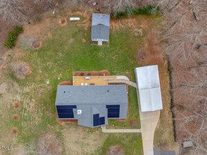 Aerial view of a property with a main house featuring a large deck and solar panels, a detached garage, and a smaller shed. The yard is spacious with scattered trees and brown grass. A driveway leads from the road to the garage.