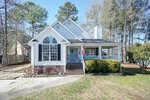 A charming white house with a dark roof and large arched window stands on 2039 Mangum Avenue in Creedmoor. An American flag hangs by the entrance. Surrounded by trees, it boasts a well-kept lawn and bushes. Steps lead to the inviting front porch, with a driveway conveniently on the left.
