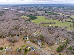 Aerial view of a rural landscape with scattered houses, winding roads, and leafless trees. Open fields and forests are visible, with a lake in the background under a cloudy sky.