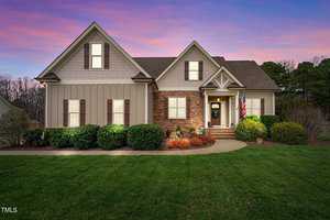 A charming house on 3853 Ironwood Drive with a mix of stone and siding under a twilight sky. It proudly displays an American flag by the entrance, while the well-maintained yard in Franklinton features lush bushes and a vibrant lawn against a sky painted in purple and pink hues.
