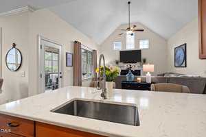 A modern kitchen with a marble countertop, sink, and faucet in the foreground. In the background, a spacious living room with a ceiling fan, TV, sofa, and a round wall mirror. French doors open to a view outside. Neutral tones create a cozy atmosphere.